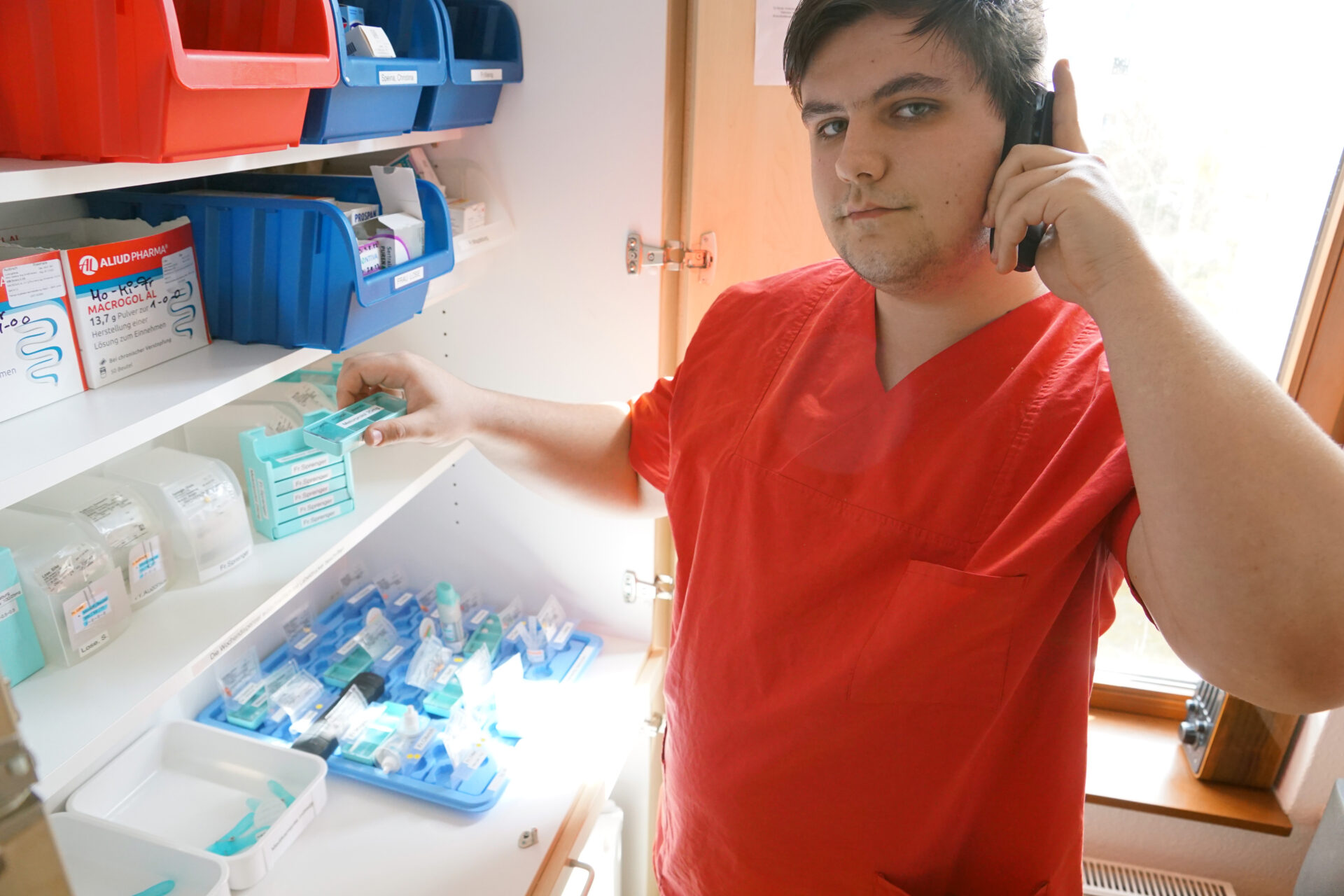 Ein junger Mann steht vor einem Medikamentenschrank und telefoniert nebenbei.