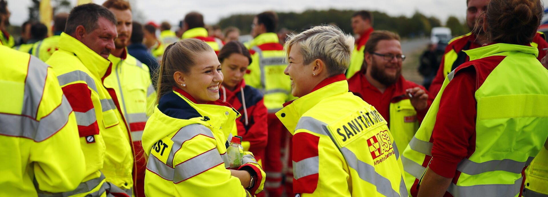 Eine große Gruppe an ASB-Sanitätern und Ehrenamtlichen sind zun einer Übung versammelt. Zwei Frauen in Warnjacken unterhalten sich freudig.