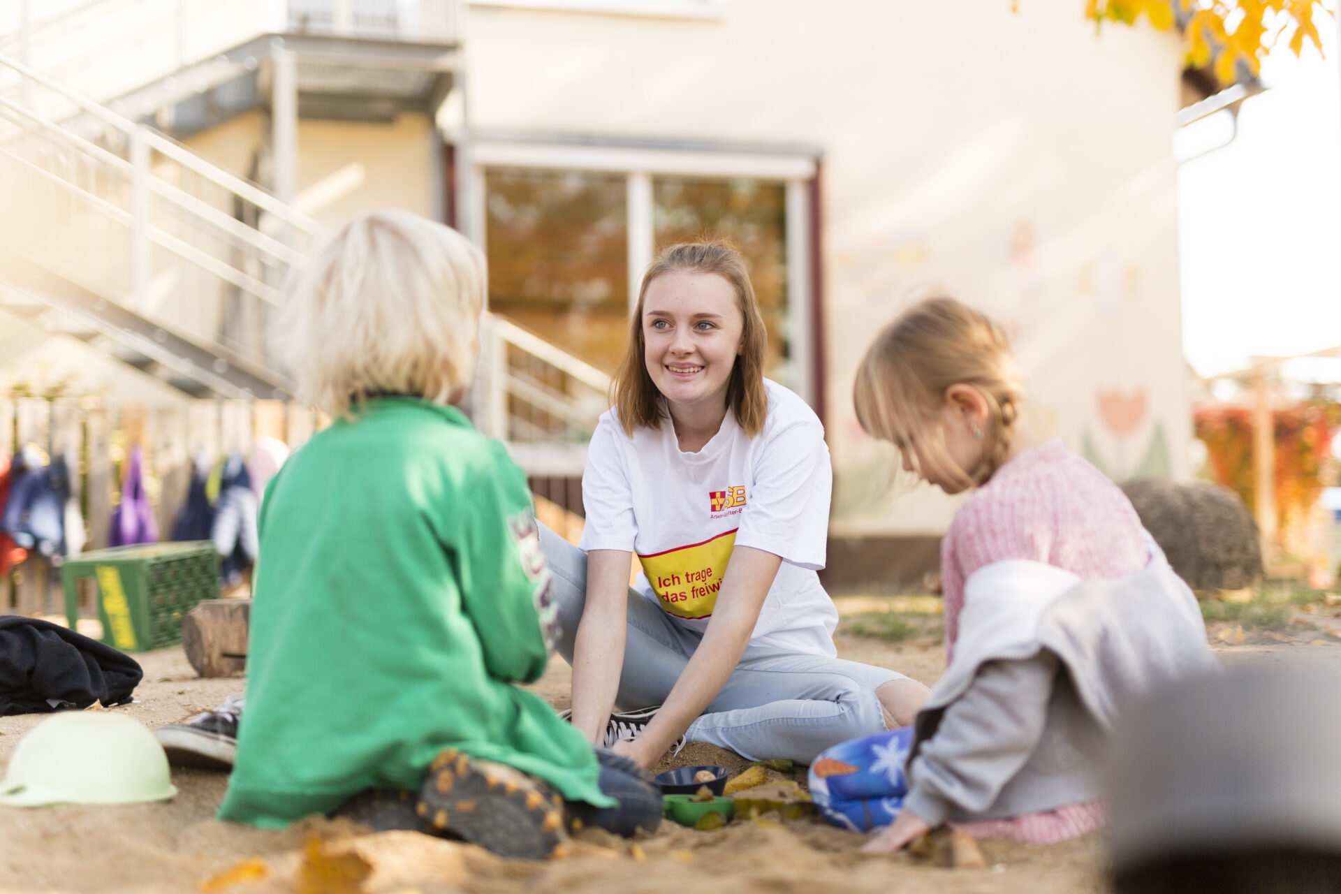 Eine junge Kindergartenerzieherin sitzt mit einem kleinen Jungen und einem kleinen Mädchen im Sandkasten.