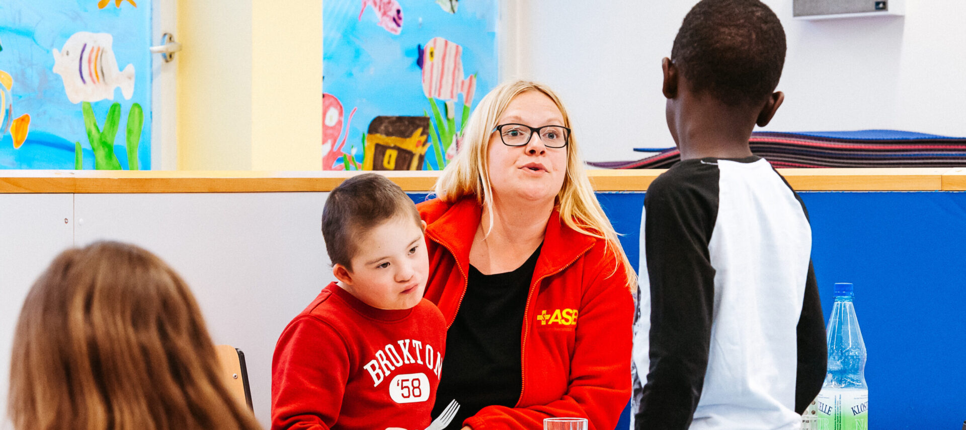 Eine Erzieherin spricht zu einem Jungen mit schwarzer Hautfarbe, während sie einem Kind mit Down-Syndrom beim Essen Gesellschaft leistet.