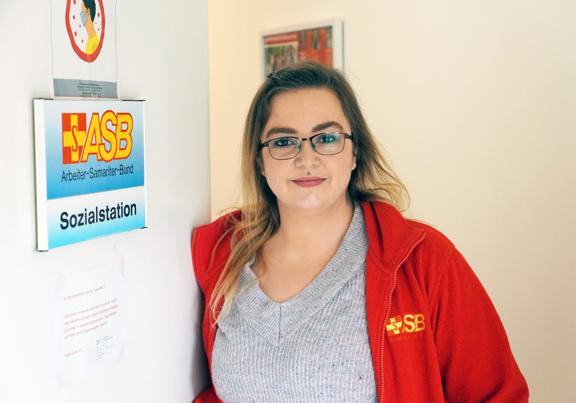 Eine junge Frau steht vor einer Sozialstation und schaut freundlich in die Kamera.