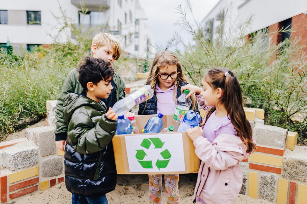 Vier Kinder halten eine Recycling-Box in der Hand und legen Plastikflaschen hinein.