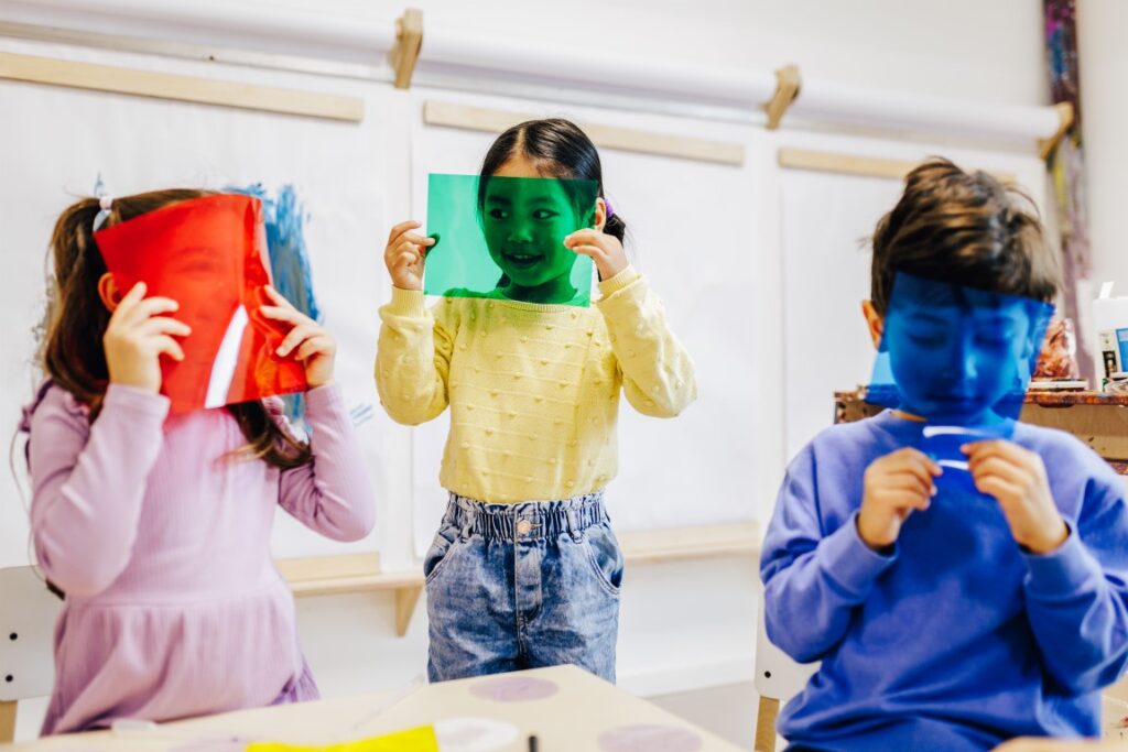 Drei Kinder halten sich jeweils eine gefärbte Plastikscheibe vor ihr Gesicht. Zwei von ihnen lachen.