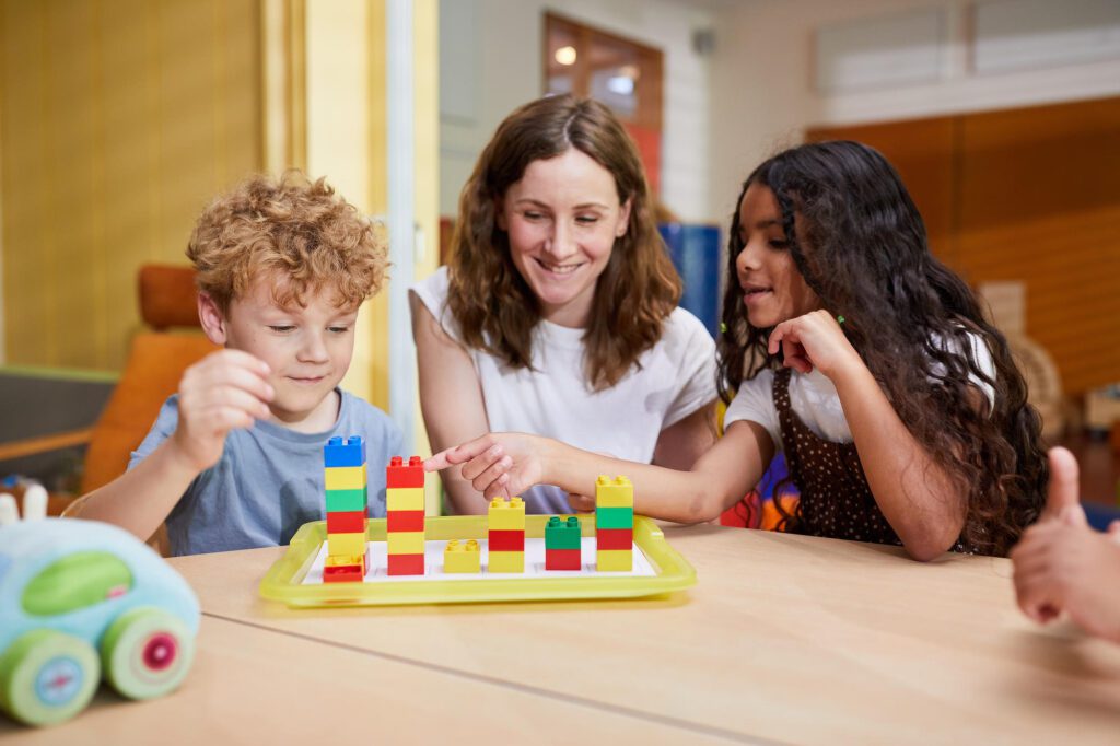 Zwei Kinder und eine Frau sitzen an einem Tisch und schauen sich Lego-Türme an, die vor ihnen stehen. Das Mädchen auf der rechten Seite zeigt auf einen der Türme.