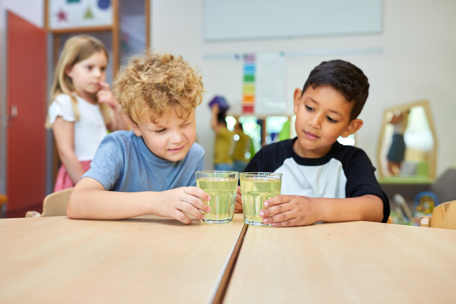 Zwei Jungen sitzen an einem Tisch und schauen sich zwei mit buntem Wasser gefüllte Gläser an. Im Hintergrund sieht man ein Mädchen und weitere Kinder.