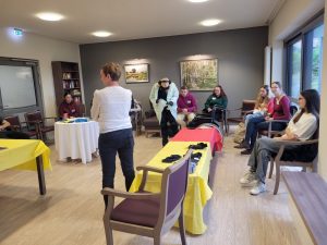 Eine Gruppe junger Menschen sitzt in einer Runde und schauen einer jungen Frau dabei zu, wie sie sich mit dem Alterssimulationsanzug versucht, einen Bademantel anzuziehen.