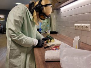 Eine junge Frau versucht konzentriert mit dem Alterssimulationsanzug eine Zucchini zu schälen.