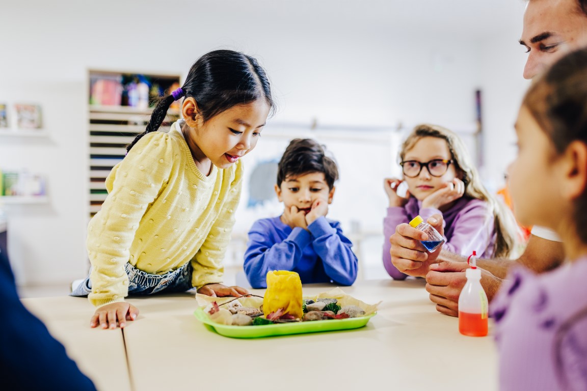 Ein paar Kinder sitzen rundherum an einem Tisch und schauen sich den in der Mitte stehenden Teller an, auf dem sich Steine, Äste und ein gelber Behälter befinden. Ein Mädchen schaut lächelnd von oben in den gelben Behälter.
