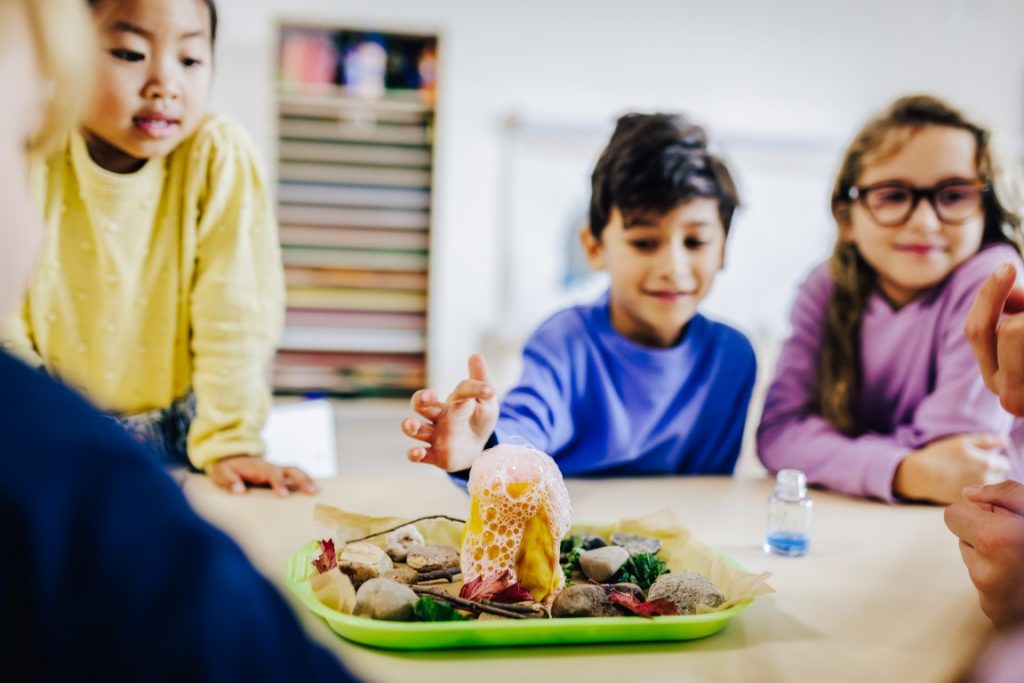 Ein paar Kinder sitzen rundherum an einem Tisch. In der Mitte des Tisches steht ein Teller, darauf Steine und Äste und ein gelber Behälter, aus dem Schaum austritt.