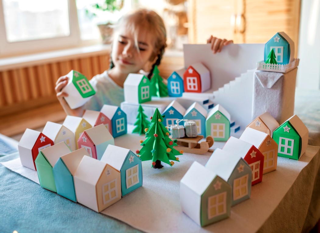Ein Mädchen sitzt im Hintergrund und schaut sich aus buntem Papier gefertigte Häuser an. Die Häuser stehen im Vordergrund und bilden eine kleine Winterlandschaft. Die Häuser sind mit Zahlen beschriftet.