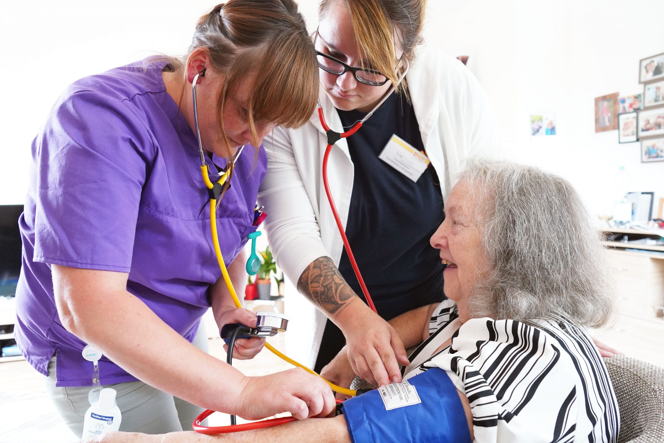 Eine alte Dame sitzt auf einem Sessel. Zwei junge Frauen stehen neben ihr und messen ihren Blutdruck.