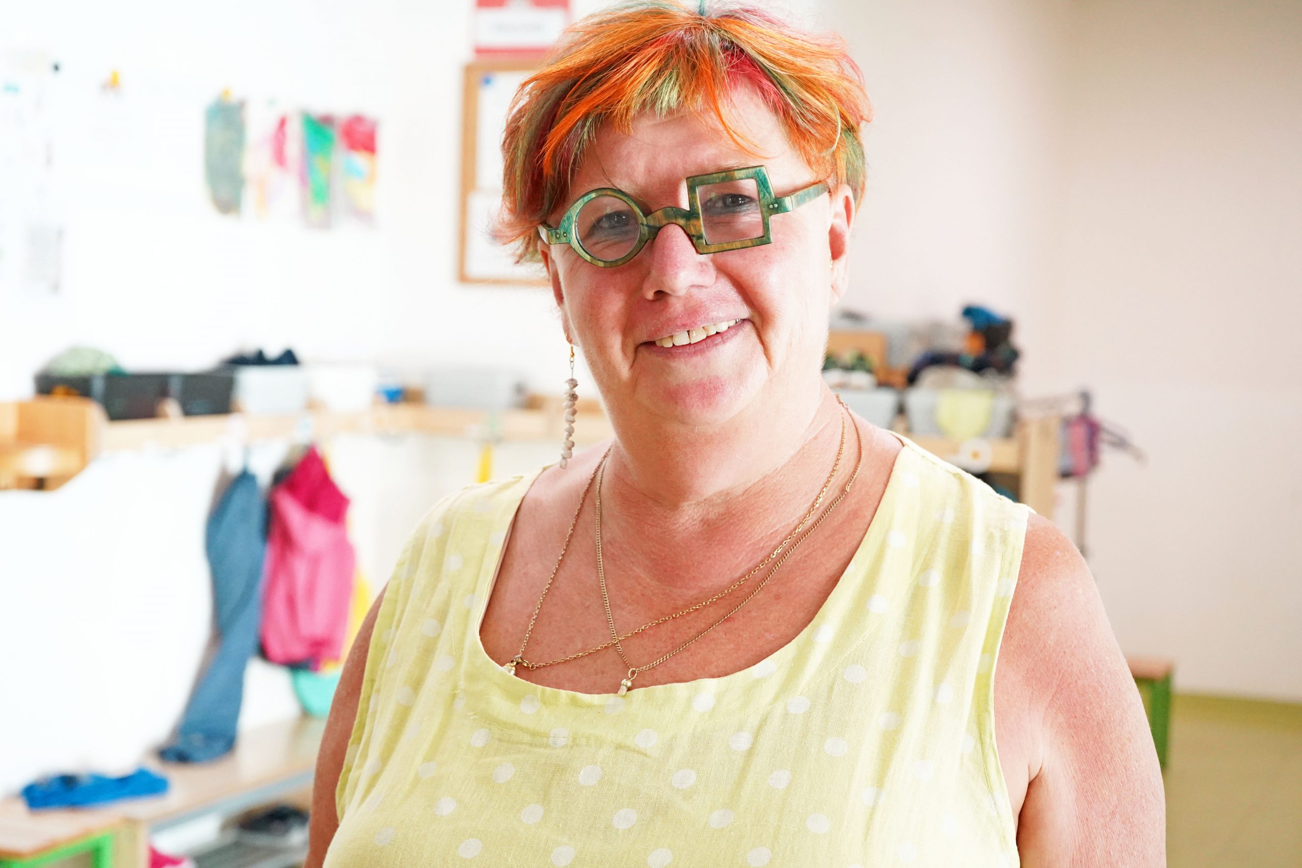 Eine ältere Dame mit bunten kurzen Haaren und einer Brille, welche zur Hälfte eckig und zur Hälfte rund ist, lächelt freundlich in die Kamera.