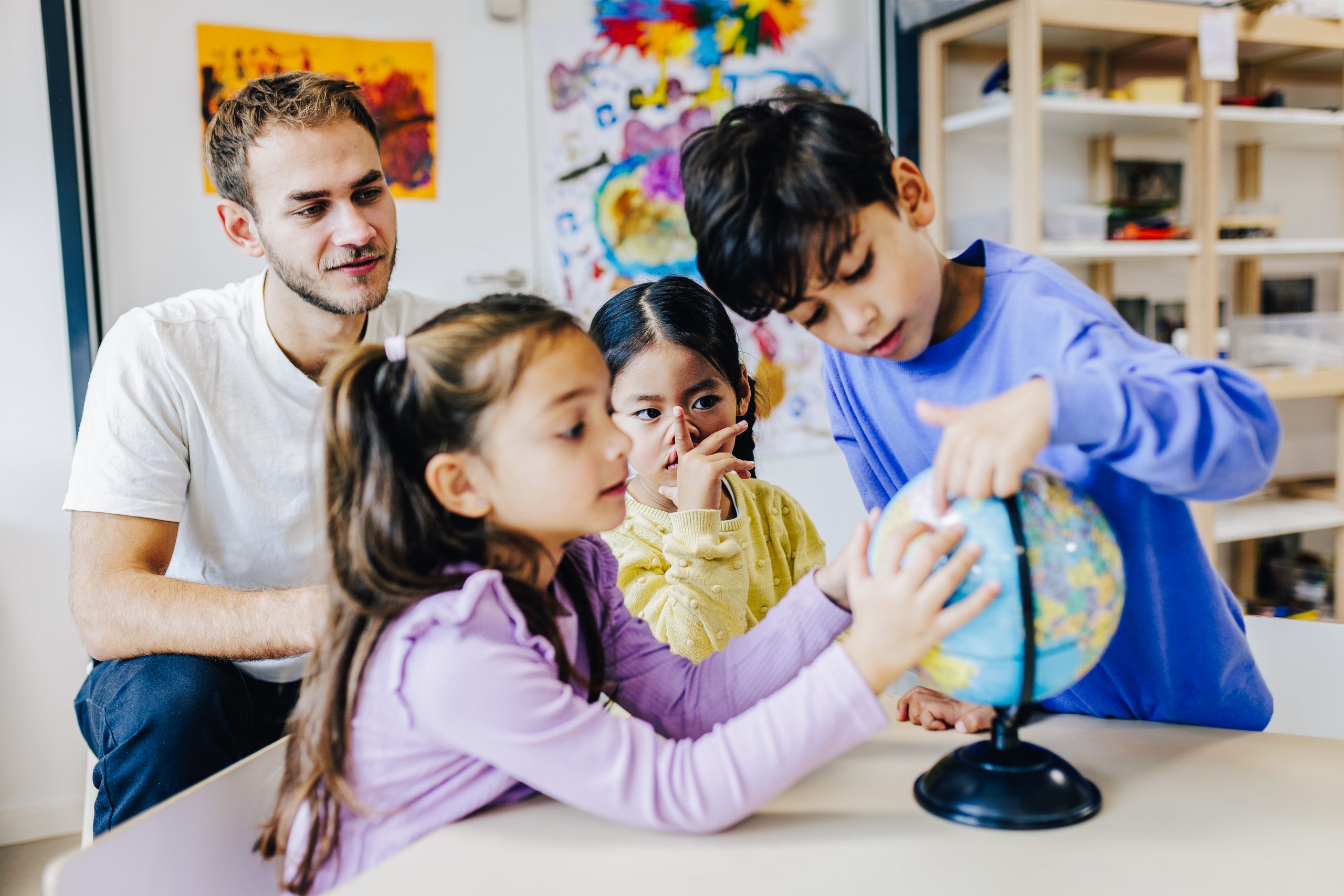 Drei Kinder schauen sich eine kleine Weltkugel an und zeigen mit ihren Fingern darauf. Ein Erzieher sitzt hinter ihnen und sieht ihnen zu.