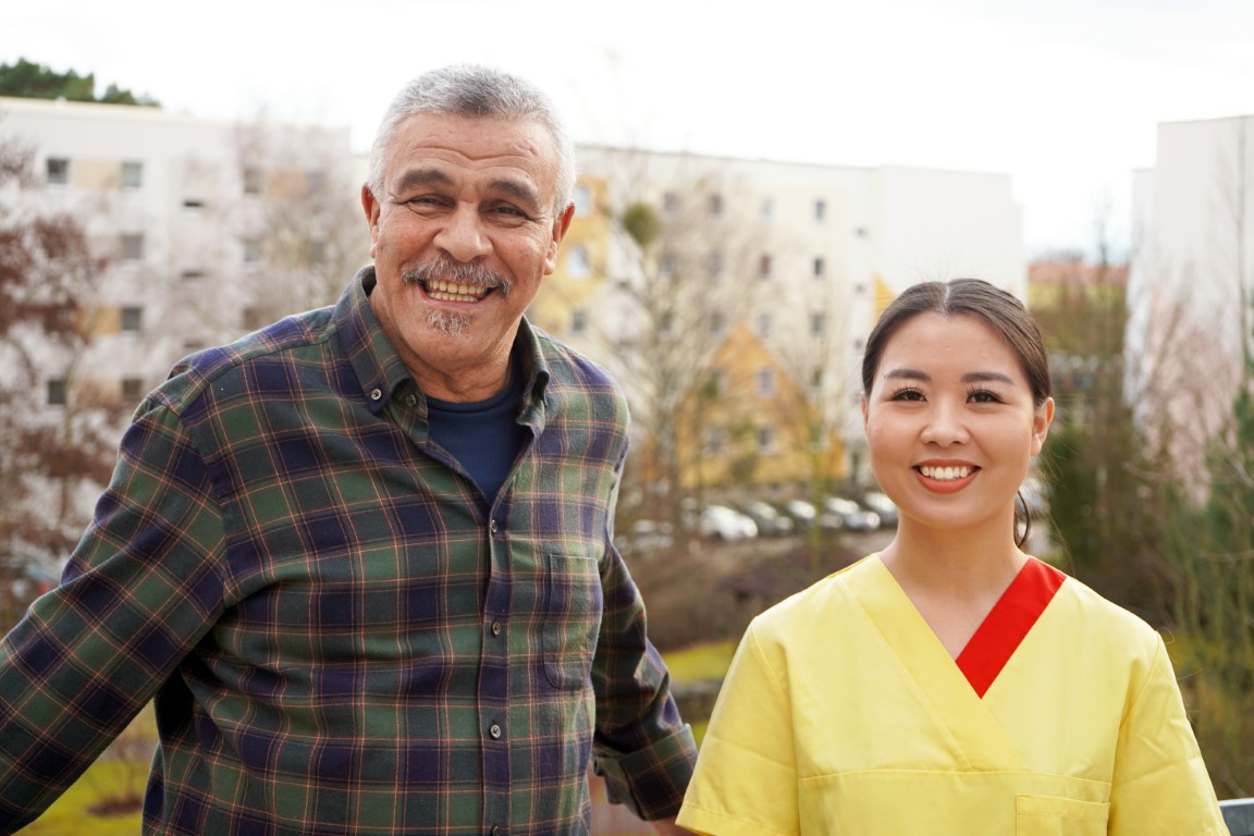 Ein älterer Herr und eine junge Dame in Pflegekleidung stehen nebeneinander und lachen freundlich in die Kamera.