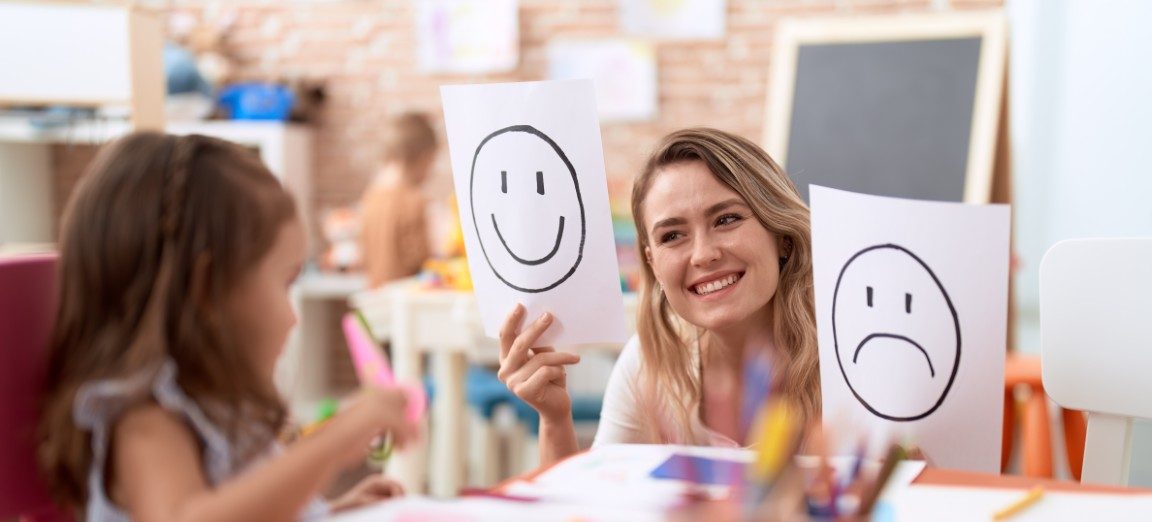 Eine junge Frau hält zwei weiße Blatt Papier in den Händen, eines mit einem glücklichen und eines mit einem traurigen Smiley und grinst dabei. Ein junges Mädchen schaut sie neugierig an.